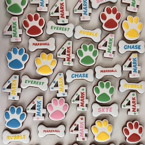 May include: A collection of decorated cookies shaped like paw prints, bones, and the number 4. The bones feature names like Rubble, Skye, Everest, Marshall, and Chase. The number 4 cookies are decorated with the name Mark and small paw prints.