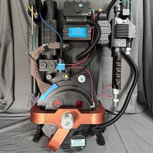 May include: A detailed prop replica of a Ghostbusters proton pack. The pack features a black and gray color scheme with copper-colored accents. It includes various tubes, wires, and a wand-like device, with warning labels. The overall design is intricate and realistic.
