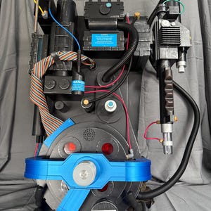 May include: A black and gray prop Ghostbusters proton pack with blue and red accents. The pack features various tubes, wires, and a blue belt-like component. The text "DANGER" is visible on a label. A prop for cosplay or display.