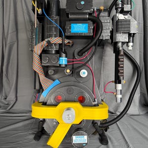 Puede incluir: Réplica detallada de un prop de una mochila de protones de Cazafantasmas. La mochila presenta una combinación de colores gris y negro con detalles amarillos, incluido un asa amarilla prominente. Tiene varios tubos, cables y un asa, con una etiqueta "DANGER".