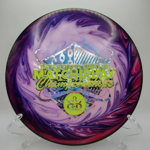 May include: A disc golf disc with a swirling pattern of purple, pink, and white. The disc features the text "UNITED STATES AMATEUR MATCHPLAY Championships" in green and yellow. A clear plastic stand supports the disc.