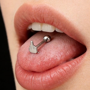 May include: A silver tongue piercing featuring a middle finger design. The barbell is inserted through the tongue, with a small ball on one end and the finger on the other. The tongue is pink, and the lips are a natural color.