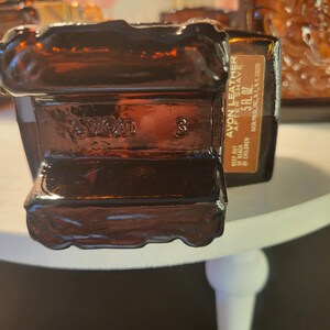 May include: A brown glass bottle of Avon Leather After Shave. The bottle is rectangular with a textured design and the text "AVON LEATHER AFTER SHAVE 5 FL. OZ." The bottle is sitting on a white surface.