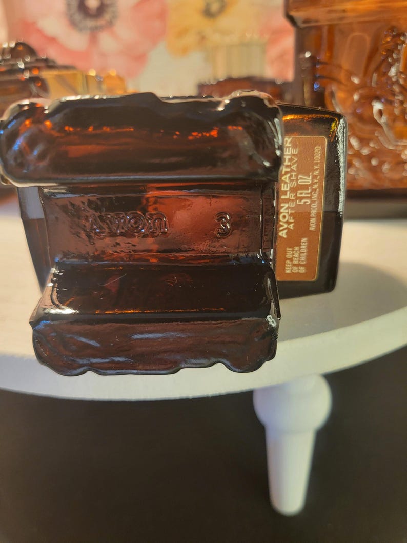 May include: A brown glass bottle of Avon Leather After Shave. The bottle is rectangular with a textured design and the text "AVON LEATHER AFTER SHAVE 5 FL. OZ." The bottle is sitting on a white surface.