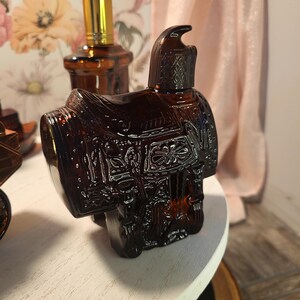 May include: A brown glass bottle shaped like a saddle, with intricate embossed details. The bottle has a dark amber hue and is positioned on a white surface. A second bottle with a gold cap is visible in the background.