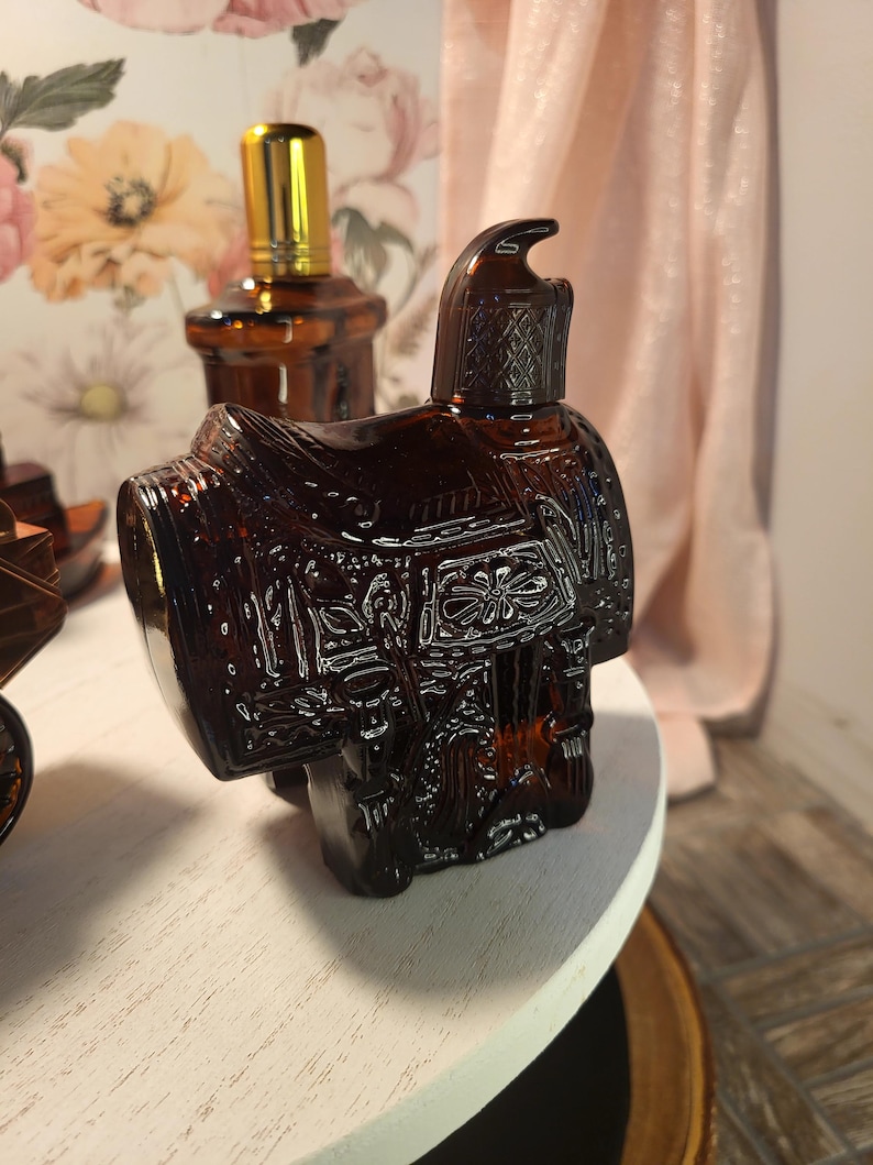 May include: A brown glass bottle shaped like a saddle, with intricate embossed details. The bottle has a dark amber hue and is positioned on a white surface. A second bottle with a gold cap is visible in the background.