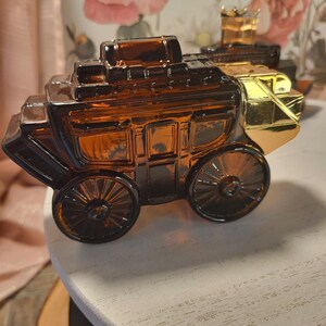 May include: Amber glass stagecoach-shaped perfume bottle with gold accents. The bottle features detailed molding of a classic stagecoach design, including wheels and a roof. A second bottle is visible in the background.