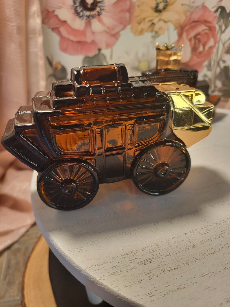 May include: Amber glass stagecoach-shaped perfume bottle with gold accents. The bottle features detailed molding of a classic stagecoach design, including wheels and a roof. A second bottle is visible in the background.