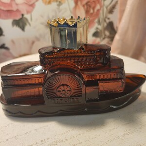 May include: A vintage amber glass steamboat-shaped bottle with a decorative crown-shaped stopper. The bottle features intricate detailing, including a paddle wheel and the word "BATCHELOR". The stopper has gold accents.