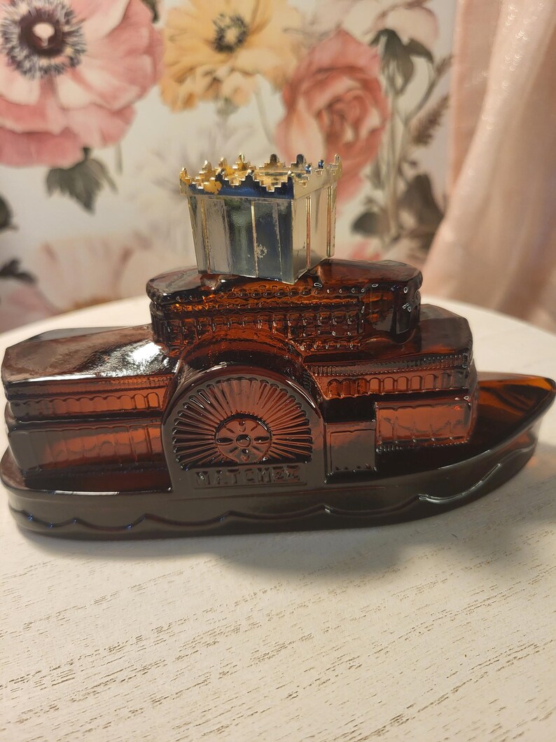 May include: A vintage amber glass steamboat-shaped bottle with a decorative crown-shaped stopper. The bottle features intricate detailing, including a paddle wheel and the word "BATCHELOR". The stopper has gold accents.