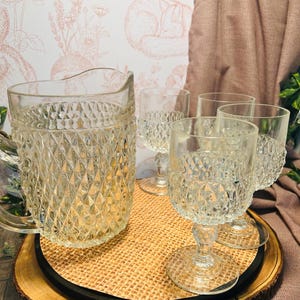 May include: A clear glass pitcher and four matching stemmed glasses with a diamond pattern. The glassware sits on a round woven tray with a black border, placed on a wooden base. The background features a floral wallpaper and a draped fabric.