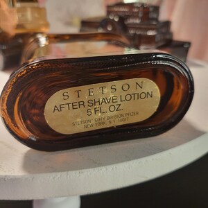 May include: A brown glass bottle of Stetson After Shave Lotion. The label is gold with black text, stating "STETSON AFTER SHAVE LOTION 5 FL. OZ." and the manufacturer's details. The bottle is oval-shaped and appears to be partially full.