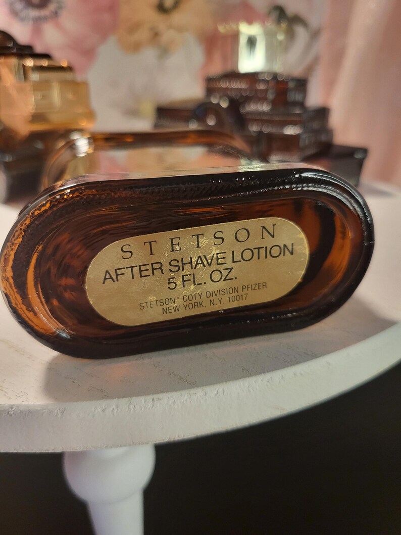 May include: A brown glass bottle of Stetson After Shave Lotion. The label is gold with black text, stating "STETSON AFTER SHAVE LOTION 5 FL. OZ." and the manufacturer's details. The bottle is oval-shaped and appears to be partially full.