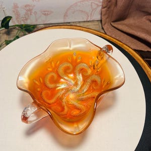 May include: An iridescent orange glass bowl with a handle. The bowl has a ruffled edge and a decorative, swirling pattern in the center. The glass has a shiny, reflective quality, and the bowl is likely vintage or antique.
