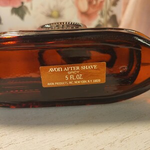 May include: A vintage Avon After Shave bottle in a rich amber color. The label reads "Spicy" and "5 FL.OZ." The bottle has a decorative silver-toned cap. The text "AVON PRODUCTS, INC, NEW YORK, N.Y. 10020" is also visible.