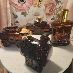May include: An assortment of vintage brown glass bottles and containers, including a stagecoach, a saddle, and a bottle with a gold cap. The items are displayed on a white surface against a floral wallpaper backdrop.