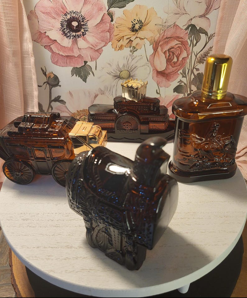 May include: An assortment of vintage brown glass bottles and containers, including a stagecoach, a saddle, and a bottle with a gold cap. The items are displayed on a white surface against a floral wallpaper backdrop.