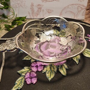 May include: A silver-colored tea strainer with a floral design. The strainer has a handle and a bowl-shaped filter with etched leaf patterns. The background features a black surface with embroidered purple flowers and green leaves.