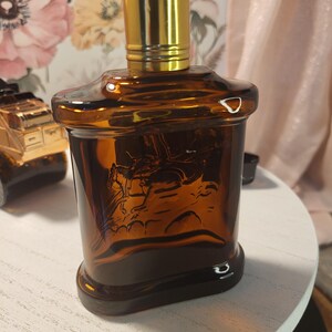 May include: A brown glass bottle with a gold-colored cap. The bottle has a rectangular shape with a detailed embossed image of a person on a horse. The bottle is sitting on a white surface.