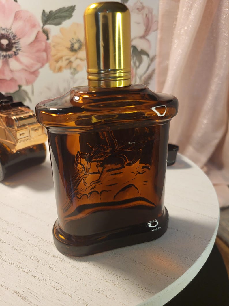 May include: A brown glass bottle with a gold-colored cap. The bottle has a rectangular shape with a detailed embossed image of a person on a horse. The bottle is sitting on a white surface.