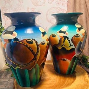 May include: Two hand-painted ceramic vases. The vases have a dark blue rim and a colorful design featuring orange, brown, and yellow shapes against a blue and green background. The vases are displayed on a wooden surface.