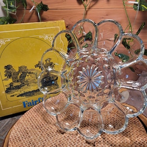May include: A clear glass serving dish with a flower-like design. The dish has a central, starburst-patterned area surrounded by petal-shaped compartments. A yellow box with a landscape illustration and the word "Fairfield" is visible in the background.
