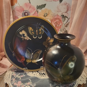 May include: A black decorative plate and vase set with butterfly designs. The plate has a gold rim and features several butterflies in shades of yellow, brown, and red. The vase is round with a narrow neck and also displays a butterfly.