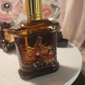 May include: A brown glass perfume bottle with a gold-colored cap. The bottle features a detailed embossed image of a person riding a horse. The bottle is sitting on a white surface.