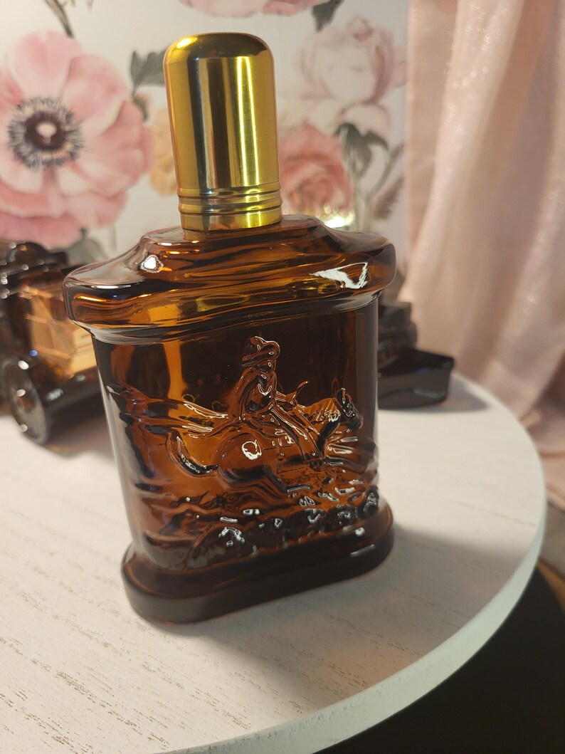 May include: A brown glass perfume bottle with a gold-colored cap. The bottle features a detailed embossed image of a person riding a horse. The bottle is sitting on a white surface.