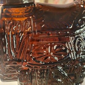 May include: A detailed close-up of a brown glass bottle with intricate embossed designs. The bottle features a decorative shape, possibly a historical figure or object, with ornate detailing and a glossy finish. The glass has a rich, amber hue.