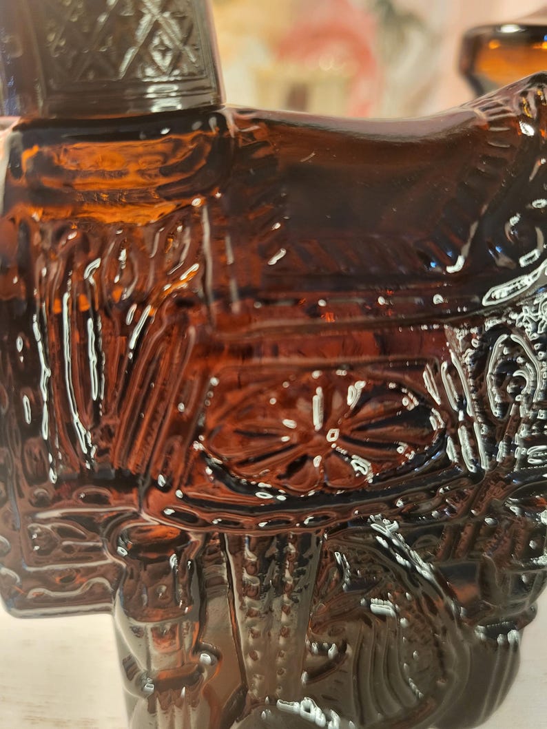 May include: A detailed close-up of a brown glass bottle with intricate embossed designs. The bottle features a decorative shape, possibly a historical figure or object, with ornate detailing and a glossy finish. The glass has a rich, amber hue.
