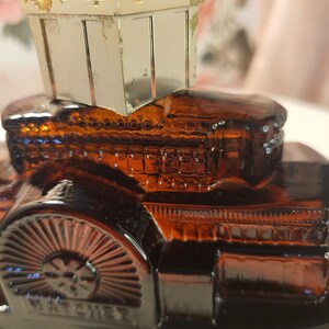 May include: An amber-colored glass car-shaped perfume bottle with a gold-colored metal top. The bottle has intricate detailing, including a wheel design and the word "NATCHEZ" embossed on the side. The top is a small, rectangular box with a crown-like design.