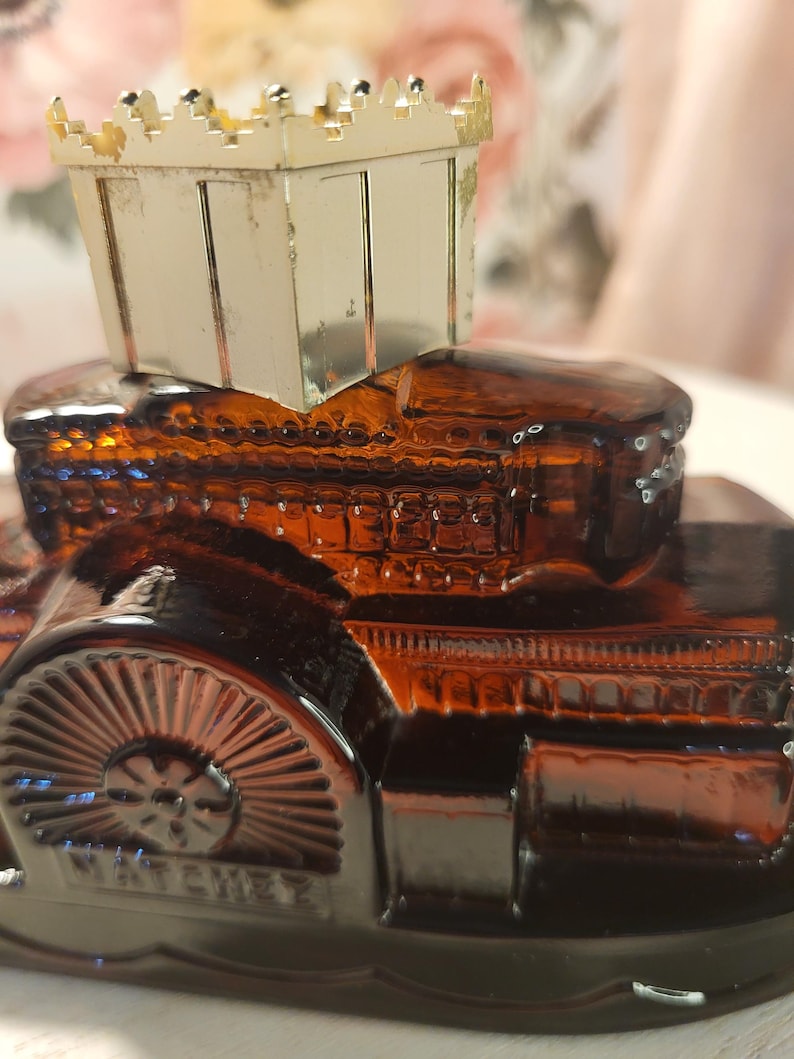 May include: An amber-colored glass car-shaped perfume bottle with a gold-colored metal top. The bottle has intricate detailing, including a wheel design and the word "NATCHEZ" embossed on the side. The top is a small, rectangular box with a crown-like design.