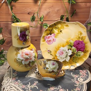 May include: A vintage tea set with a yellow background and floral rose design. The set includes a teacup, saucer, and a two-piece decorative item. The roses are in shades of pink, purple, and white.