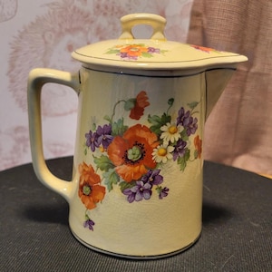 May include: A vintage cream-colored ceramic pitcher with a lid, adorned with a floral design featuring orange poppies, purple flowers, and white daisies. The pitcher has a handle and a spout, and the lid has a small handle.