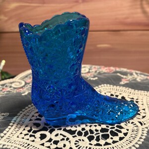 May include: A vibrant blue glass boot-shaped vase with a textured floral pattern. The vase has a scalloped top and is likely vintage. The boot is a decorative item, possibly for small flowers or as a collectible.