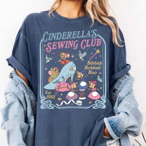 May include: A navy blue t-shirt featuring the text "Cinderella's Sewing Club" with a whimsical illustration. The design includes Cinderella's glass slipper, mice, and the phrase "Bibbidi Bobbidi Boo." The shirt is dated "Est. 1950."