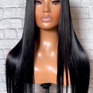 May include: A mannequin head showcasing a long, straight, black wig. The wig has a glossy, smooth texture and reaches past the shoulders. The mannequin has a tanned complexion and is set against a white brick wall.