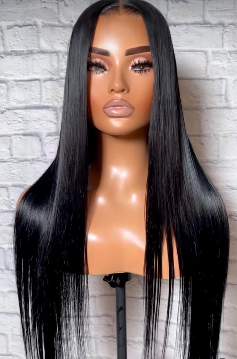 May include: A mannequin head showcasing a long, straight, black wig. The wig has a glossy, smooth texture and reaches past the shoulders. The mannequin has a tanned complexion and is set against a white brick wall.