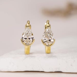 Puede incluir: Un par de pendientes de aro dorados, cada uno con una piedra preciosa transparente en forma de pera engastada en un marco de oro pulido. Los pendientes tienen un mecanismo de cierre de bisagra y un acabado metálico liso y reflectante.