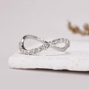 Puede incluir: Un anillo de banda curva en tono plateado con una serie de pequeñas piedras redondas y transparentes talladas a lo largo del borde superior. El metal tiene un acabado pulido y reflectante, con un diseño en forma de onda suave.