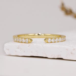 Peut inclure: Une bague ouverte en or ornée d'une rangée de petites pierres transparentes taillées en rond sur la bande avant. Le métal présente une finition polie et le design comporte un espace au centre, mettant en valeur une ligne continue de pierres.