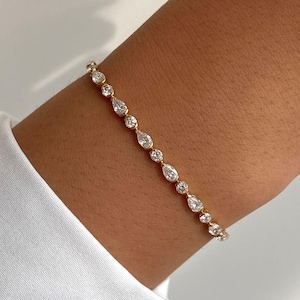 May include: A gold tennis bracelet featuring an alternating pattern of round-cut and pear-shaped clear stones. The delicate chain is crafted from yellow gold, with each gemstone securely set in a prong setting, creating a continuous line of sparkle.