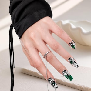 May include: Silver nail rings and nail art with green detailing. The rings have complex designs and are attached to the nails. The nails are painted green and have silver embellishments.