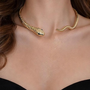 May include: A gold-coloured snake necklace with a textured design. The snake's head is at the front, and the tail curves around the neck. The necklace is worn over a black strapless top. A heart-shaped brooch with a large yellow stone is also visible.