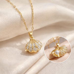 Puede incluir: Un collar dorado con un colgante de concha adornado con cristales transparentes. El colgante cuelga de una delicada cadena. Una pequeña incrustación circular muestra el colgante con una perla en su interior.
