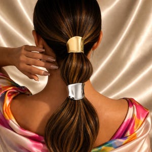 Puede incluir: Peinado de cola de caballo con dos brazaletes de pelo metálicos. El brazalete superior es dorado y el inferior plateado. El cabello es castaño con reflejos. El fondo es de tela satinada.