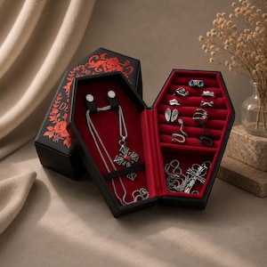 May include: Black coffin-shaped jewelry box with red velvet interior, displaying a silver necklace with a cross pendant and various rings. The box is open, revealing the jewelry. The exterior is black with red floral and skull designs.