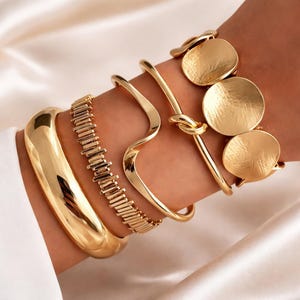 May include: A selection of gold-coloured bracelets. The bracelets include a thick, rounded bangle, a rectangular link bracelet, a wavy design, a knot design, and a bracelet with three large, round, textured charms.