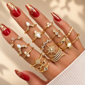 Op de afbeelding: Een hand versierd met meerdere gouden ringen met verschillende ontwerpen, waaronder opalen, parels en geometrische vormen. De ringen zijn in verschillende stijlen, zoals banden, midi-ringen en statement-stukken. De nagels zijn rood geverfd met gouden glitter.